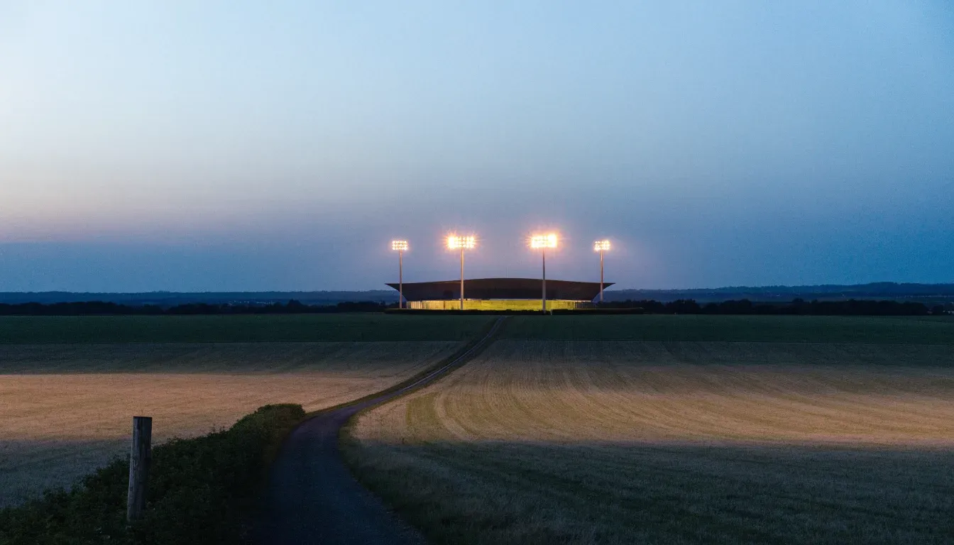 Ein Stadion unter Flutlicht aus großer Entfernung, davor Felder und eine ruhige Straße in der Dämmerung.