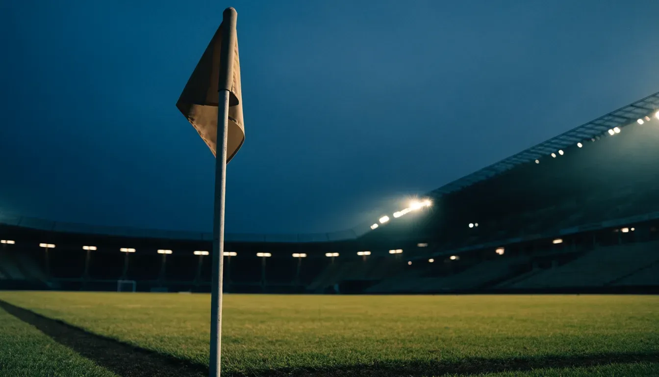 Eine Eckfahne am Rand eines Rasenplatzes, dahinter ein leeres Stadion unter Flutlicht aus der Ferne gesehen.